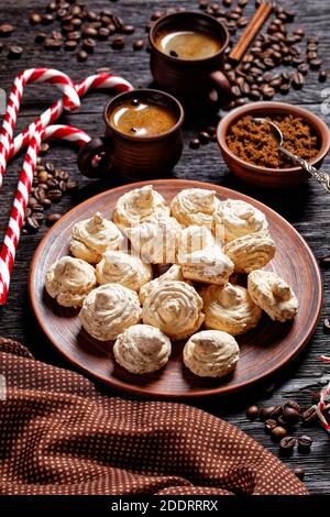 Biscotti meringa serviti su un piatto con canna da zucchero di natale, tazze di caffè, chicchi di caffè, zucchero di canna e bastoncini di cannella su uno sfondo di legno scuro, Foto Stock