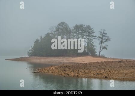 Fitta nebbia mattutina al lago con i boschi su una piccola isola oscurata dalle condizioni del tempo in inverno Foto Stock