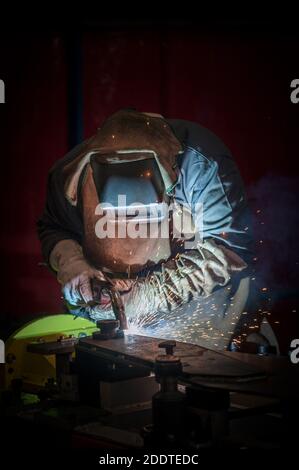 Saldatrice al lavoro in una fabbrica con attrezzatura di protezione completa - dispositivi di protezione individuale (DPI) - su sfondo rosso scuro Foto Stock