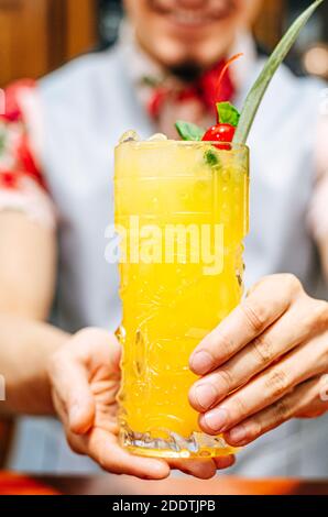 Barista che mostra un cocktail in un bicchiere di tiki su un bar contatore su uno sfondo sfocato di bottiglie in una barra Foto Stock