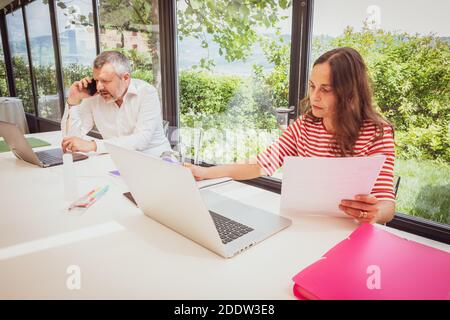 L'uomo impegnato e serio sta lavorando con il suo laptop mentre la donna lavora nelle vicinanze a casa - madre e padre lavorano da remoto da casa Foto Stock