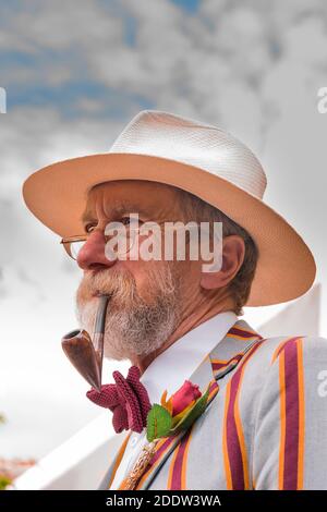 Primo piano ritratto di senior gentleman in estate paglia trilby cappello, fumo di una pipa, indossando intelligente barca blazer, fiore in lapel. dapper gent isolato. Foto Stock