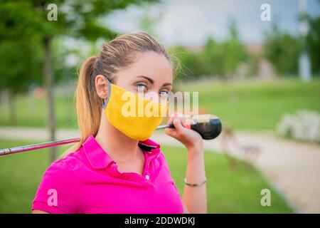 Ritratto di una bella donna bionda che sta giocando a golf su sfondo di campo verde all'aperto con maschere protettive - giovane ragazza con golf club sti Foto Stock