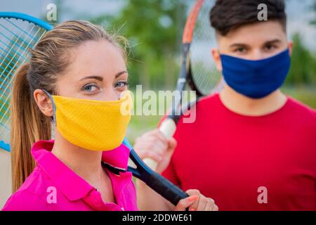 Ritratto di tennisti che tengono la racchetta all'esterno con maschere protettive - primo piano ragazza bionda e ragazzo che sono giocare a tennis durante la quarantena Foto Stock