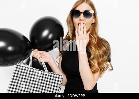 Una ragazza felice scioccata in occhiali da sole, con borse e palloncini, copre la sua bocca con la mano in delizia, su uno sfondo bianco . Shopping, discoun Foto Stock