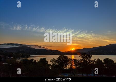 Alba su montagne e lago, vicino alla città costiera. Foschia di nebbia sulle montagne, il riflesso del sole nell'acqua, sagome degli alberi i Foto Stock