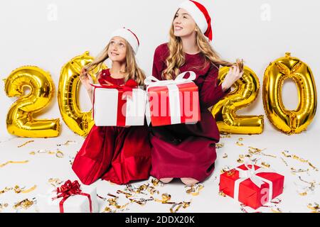 Una giovane famiglia, madre e bambina a Santa cappelli, seduta pensierosa su uno sfondo bianco con regali di Natale in mano, con palloncino Foto Stock