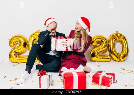 Una giovane famiglia di Natale, un ragazzo e una ragazza in cappelli di Santa, danno regali seduti su uno sfondo bianco con palloncini sotto forma di figure 2020 Foto Stock