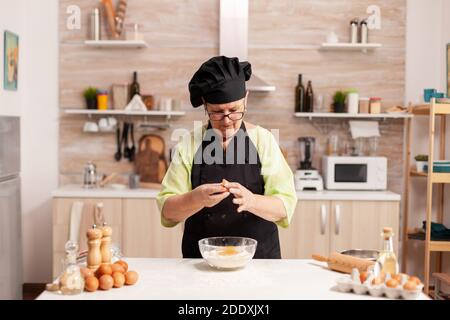 Mani dello chef con farina e uova nel processo di preparazione per la cottura. Cuoco di pasta anziano che coglie l'uovo sulla ciotola di vetro per la ricetta della torta in cucina, mescolando a mano, impastando. Foto Stock