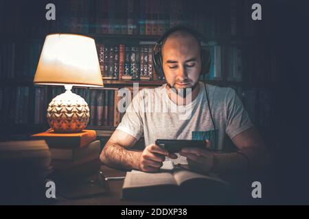 Lettura ed educazione. Un uomo della biblioteca sta ascoltando un audiolibro, o sta guardando un video sul telefono, e c'è un libro aperto sul tavolo. Poll Foto Stock