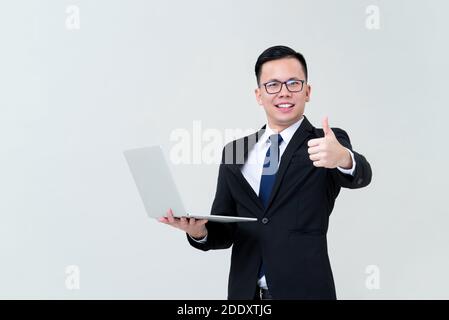 Giovane uomo d'affari asiatico essere felice con il suo commercio in linea che trasporta laptop e dando pollici isolati su sfondo grigio studio Foto Stock