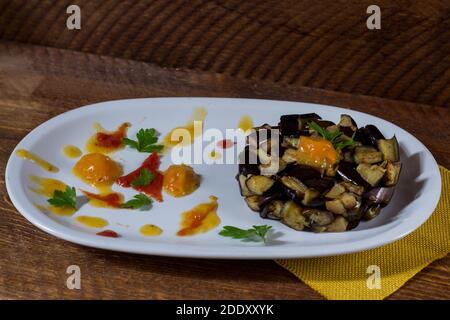 Piatto di melanzane a dadini su una crema di pomodori ciliegini gialli e rossi in un piatto bianco. Legno di fondo. Messa a fuoco selettiva. Foto Stock