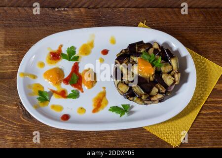 Piatto di melanzane a dadini su una crema di pomodori ciliegini gialli e rossi in un piatto bianco. Legno di fondo. Messa a fuoco selettiva. Foto Stock