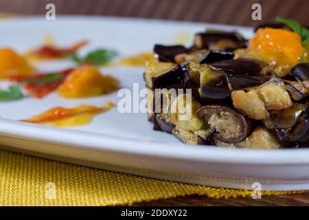 Piatto di melanzane a dadini su una crema di pomodori ciliegini gialli e rossi in un piatto bianco. Legno di fondo. Messa a fuoco selettiva. Foto Stock