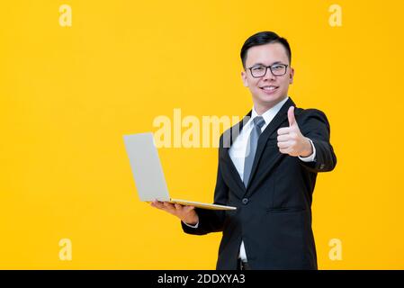 Giovane uomo d'affari asiatico essere felice con il suo commercio in linea che trasporta computer portatile e dando pollici isolati su studio giallo sfondo Foto Stock