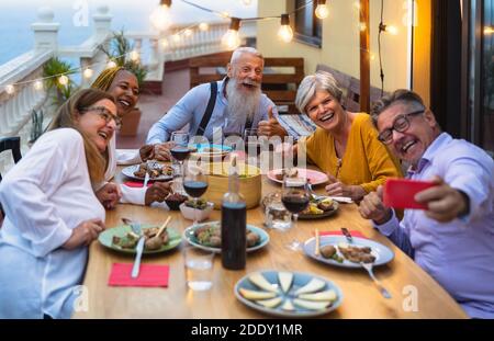 Amici anziani multirazziali felici di divertirsi a cenare insieme mentre si prende selfie con smartphone mobile sul patio della casa Foto Stock
