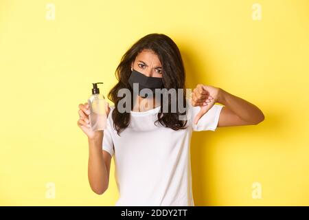 Concetto di covid-19, divaricamento sociale e stile di vita. Immagine della donna afro-americana delusa in maschera facciale, mostrando pollici-giù, non raccomandare Foto Stock