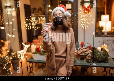 Giovane donna felice in maschera facciale che festeggia da sola le vacanze di Capodanno a casa. Concetto di quarantena e autoisolamento durante l'epidemia in vacanza Foto Stock