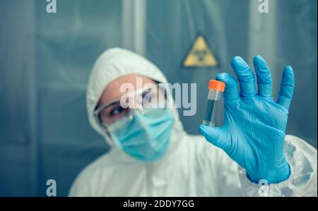 Donna scienziata con tuta di protezione batteriologica che guarda il flaconcino in laboratorio. Fuoco selettivo sul flaconcino in primo piano Foto Stock