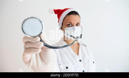 Medico femminile con un cappello di Santa e una maschera protettiva sul viso utilizzando stetoscopio isolato su sfondo bianco. Foto Stock