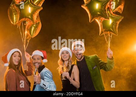 sorridenti amici multiculturali in cappelli santa con bicchieri e champagne vicino palloncini su nero Foto Stock