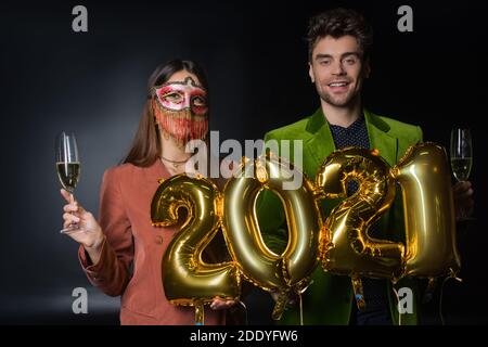 uomo felice tenendo palloncini con 2021 numeri vicino donna dentro maschera di carnevale con bicchiere di champagne su nero Foto Stock