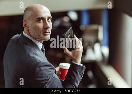 Uomo d'affari calvo che guarda la macchina fotografica girata da dietro. Uomo caucasico che indossa un vestito da lavoro, occhiali e cravatta che parla sul telefono cellulare mentre si scende Foto Stock