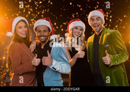 sorridendo amici interrazziali in santa cappelli mostrando pollici vicino caduta di confetti su nero Foto Stock