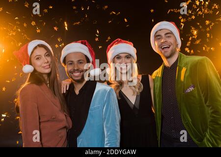 felici amici interrazziali a santa cappelli sorridenti vicino a cadere confetti su nero Foto Stock