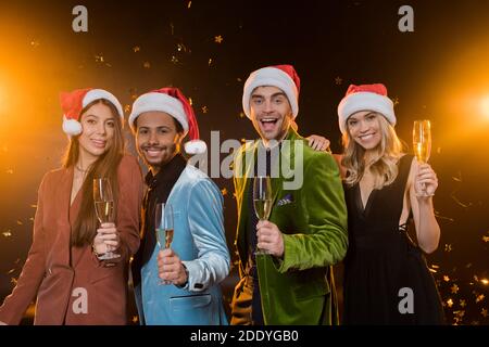 sorridenti amici interrazziali in cappelli santa con bicchieri e champagne vicino a cadere confetti su nero Foto Stock