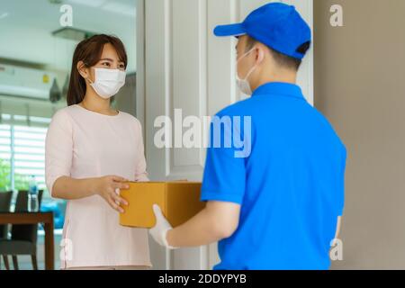Asian consegna uomo che indossa maschera facciale e guanto in uniforme blu tenendo una scatola di cartone in casa di fronte e donna che accetta una consegna di scatole da d Foto Stock