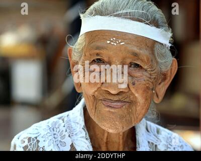 La vecchia donna balinese indonesiana con grani di riso sulla fronte indossa una blusa tradizionale bianca kebaya durante una cerimonia religiosa indù. Foto Stock