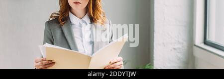 Vista ritagliata di donna d'affari redhead che tiene la cartella con fogli di carta vicino alla finestra, banner Foto Stock