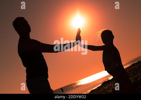 silhouette di una bambina con suo padre, palme di mani sollevate l'una verso l'altra sullo sfondo di un tramonto arancione al mare. Buon sume Foto Stock