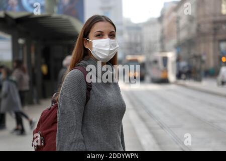 Studentessa con maschera protettiva in attesa di trasporto pubblico in strada. Giovane donna con maschera chirurgica in attesa di tram alla fermata nella città grigia inquinata. Foto Stock