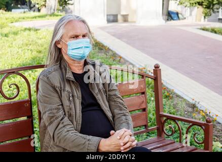 l'uomo anziano con lunghi capelli grigi in maschera protettiva si siede su una panchina in un parco cittadino in autunno soleggiato giorno Foto Stock