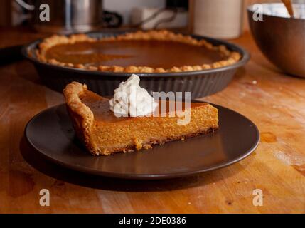Una fetta di torta di zucca fatta in casa con un dollop di panna montata si siede su un piatto su un bancone di legno, con il resto della torta sullo sfondo. Foto Stock