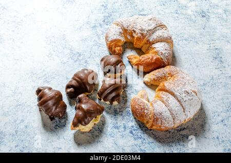Due croissant appena sfornati con zucchero di vetro e diversi mini croissant al cioccolato su sfondo blu con consistenza. Foto Stock