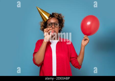 Donna africana sbalordita che soffia il salone dell'aria per la festa di compleanno. Studio girato su parete blu. Foto Stock
