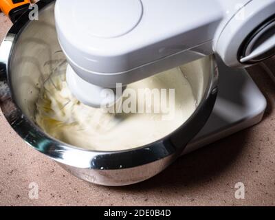 cottura dolce pan di spagna a casa - sopra la vista di mescolando la crema liquida con lo zucchero in una ciotola di acciaio di robot da cucina a casa (mixer whisk blu Foto Stock