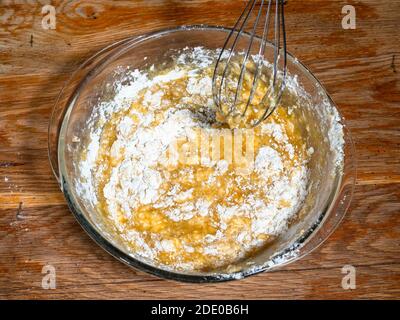 cottura dolce pan di spagna a casa - sopra la vista di mescolando l'impasto con la farina e gli ingredienti dolci con la frusta in una ciotola di vetro sul vecchio tavolo di legno a casa Foto Stock