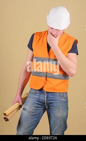 Ingegnere, architetto, operaio, costruttore sul volto premuroso tiene in mano vecchi progetti. Uomo, caposquadra in casco, elmetto ha dubbi circa la pianta della costruzione, sfondo ocra. Concetto di ingegneria. Foto Stock