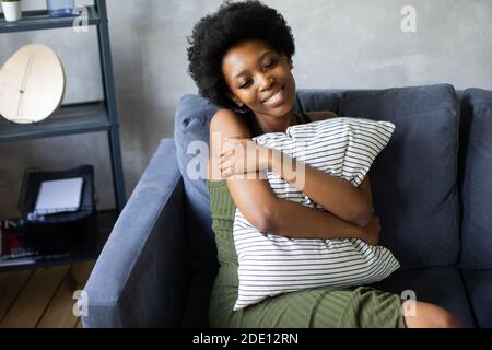 Felice african American giovane donna sedersi rilassarsi su un divano accogliente e abbracci un cuscino. Felice di trasferirsi in un nuovo appartamento. Foto Stock