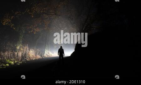 Una figura con cappuccio e una luce brillante su una strada di campagna. In una notte di inverni nebbiosa. Foto Stock