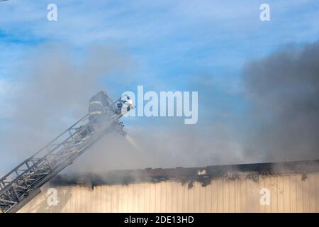 Vigili del fuoco che combattono un incendio da costruzione in cima a una scala estesa. Il fumo proveniente dall'edificio oscura parzialmente i vigili del fuoco. Foto Stock