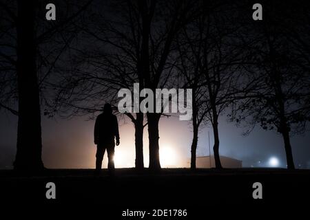 Una figura con cappuccio accanto agli alberi, con silhouette che si contrappone a una luce brillante e brillante. In una notte di inverni nebbiosa. Foto Stock