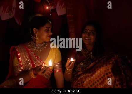 Due belle donne indiane bengalesi in abito tradizionale indiano che illumina le lampade Diwali seduta sul pavimento in serata Diwali. Stile di vita indiano Foto Stock