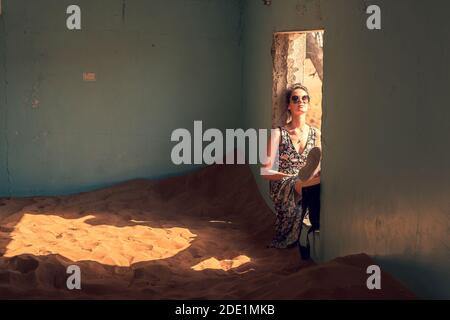 Donna in un abito colorato estivo con occhiali da sole seduti la finestra della casa abbandonata piena di sabbia in il deserto in attesa del tramonto Foto Stock