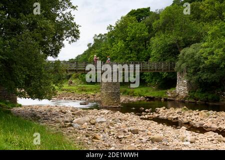 Passerella sul fiume Swale (campagna panoramica, acque basse e poco profonde, rocce fluviali, 2 persone che scattano foto) - Ramps Holme Bridge, Yorkshire Dales, Regno Unito. Foto Stock