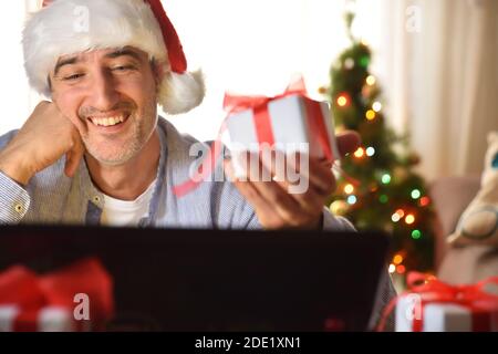 Uomo ridente con cappello di Babbo Natale e regalo in mano guardando un computer portatile nel soggiorno. Foto Stock